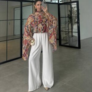 Multicolored patchwork-print poncho with scarf neckline and wide draped sleeves, paired with white wide-leg trousers.
