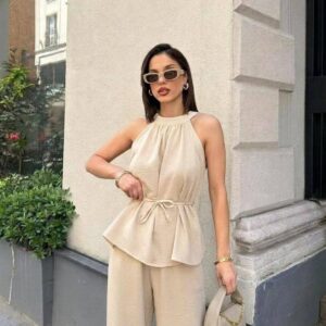 Outdoor view of beige halter peplum top with double-layered peplum and wide-leg pants, model holding a beige handbag.