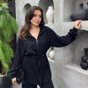 Chaqueta negra con cremallera y tirador circular, mangas sueltas y cintura anudada con cinturón, sosteniendo un clutch brillante. Disponible para mayoristas en Turquía - Mayorista Turquía.