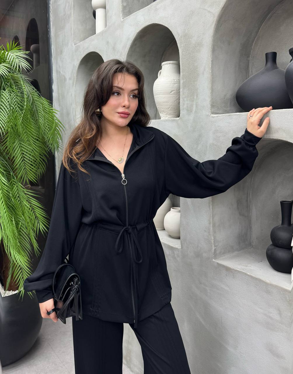negro-zip-cierre-redondo-9043 – Mayorista Turquía Chaqueta negra con cremallera y tirador circular, mangas sueltas y cintura anudada con cinturón, sosteniendo un clutch brillante. Disponible para mayoristas en Turquía - Mayorista Turquía.