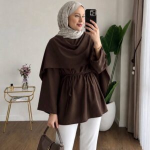 Vista frontal de tunica marrón chocolate con capa superpuesta y lazo en la cintura, bolso de cuero marrón a juego y pantalones blancos.