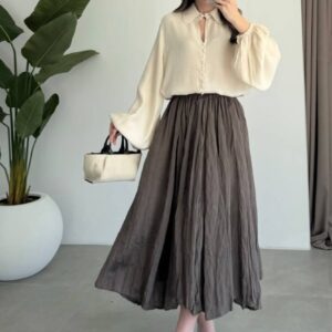Model wearing cream balloon-sleeve blouse with fabric buttons and dark brown crinkled midi skirt, holding small handbag.