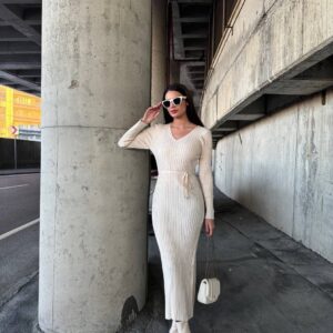 Model trägt cremefarbenes geripptes Strick-Midikleid mit V-Ausschnitt und dünnem Bindeband, kombiniert mit weißen Stiefeln und Sonnenbrille.