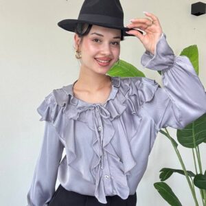Model wearing grey ruffled blouse with a black fedora, showing neckline ruffles and gathered cuffs.