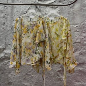 Two sheer floral chiffon blouses on white hangers showing soft yellow and purple botanical prints with asymmetrical hems and ties.
