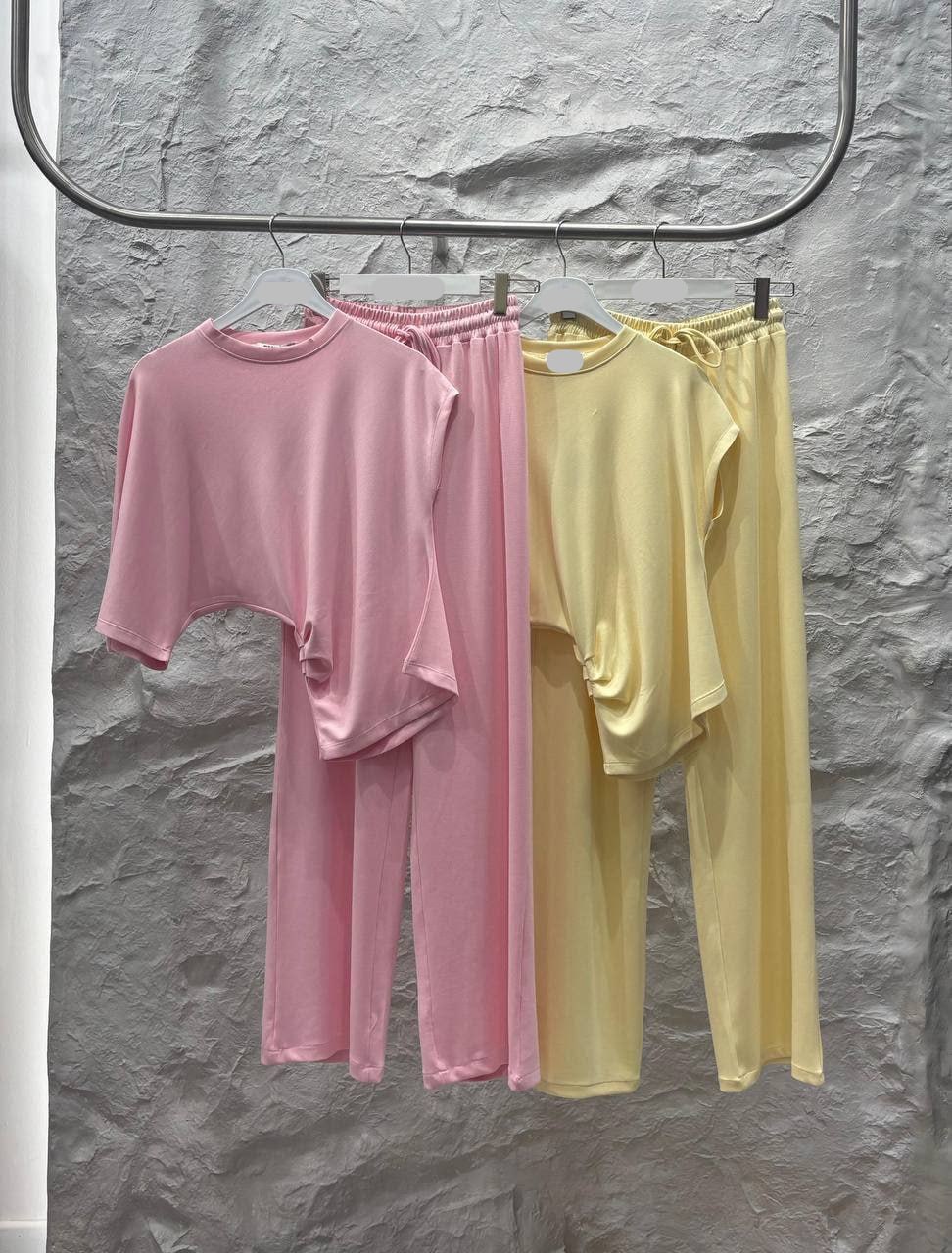 Pastell-Loungewear-Sets 3322 – Türkei Großhandel Zwei Pastell-Loungewear-Sets in Rosa und Gelb hängen an einem silbernen Regal; Oberteile und passende weite Hosen sind sichtbar. Türkei-Großhandel.