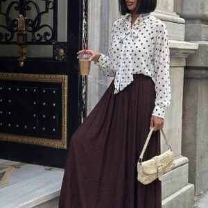 Three-quarter view of model wearing a white polka-dot tie-neck blouse and flowing brown maxi skirt, holding a beige bag and iced coffee. available for wholesale in Turkey.