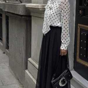 Seitenansicht einer weissen Polka-Dot-Krawattenbluse und eines schwarzen, transparent geschichteten Maxirocks; ergänzt durch schwarze Schultertasche und spitze Absätze. - Grosshandel-Tuerkei