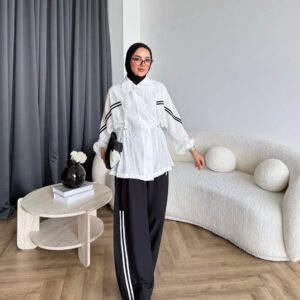 عارضة ترتدي جاكيت أبيض بسحاب مع خطوط سوداء ثلاث على الأكمام، خصر بسحاب، بنطال واسع أسود وأحذية رياضية بيضاء. متاح بالجملة من تركيا.