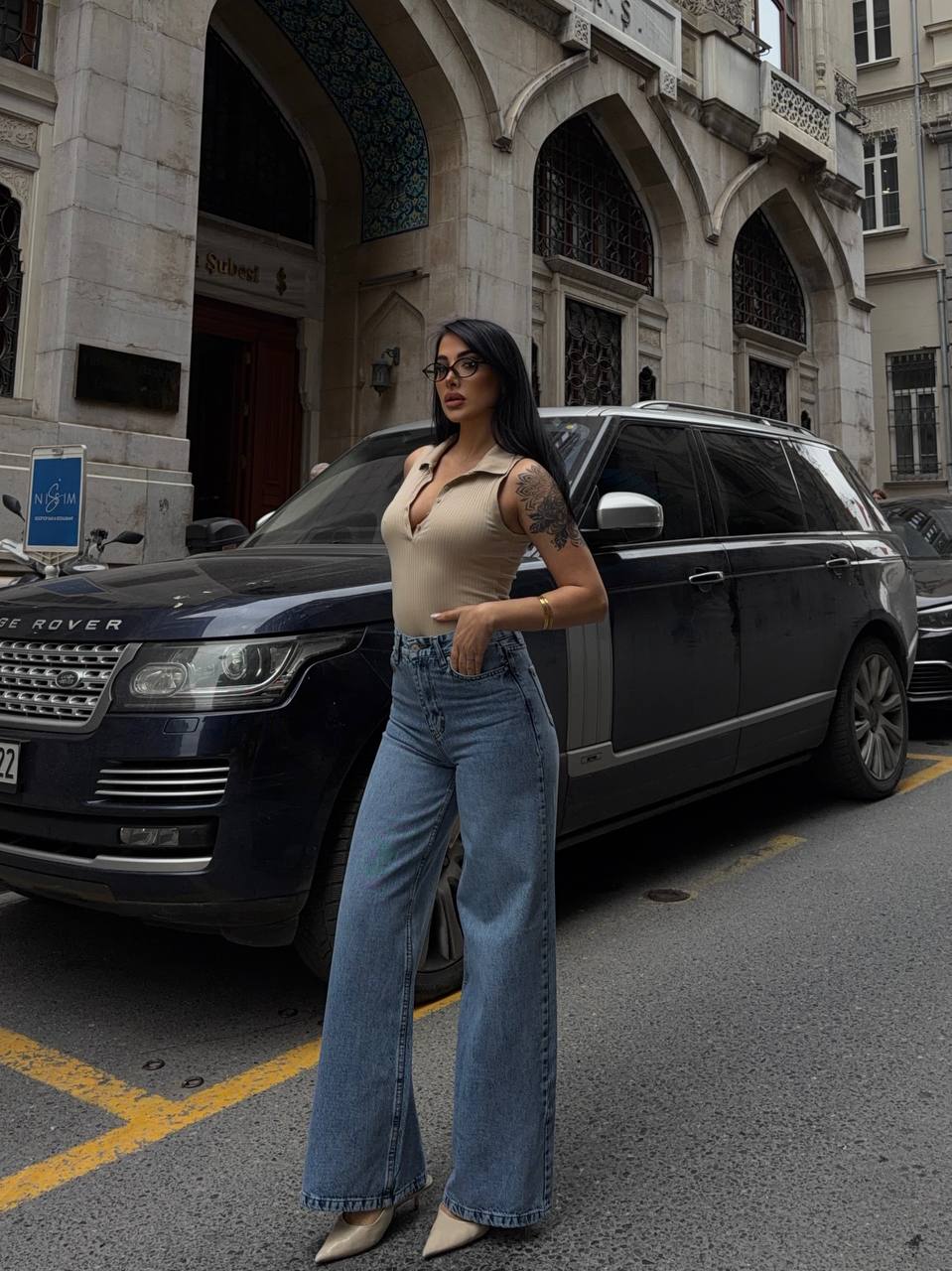 جينز دنيم واسع الساق – ستايل شارع – تركيا بالجملة امرأة تقف بجانب سيارة SUV فاخرة داكنة اللون وهي ترتدي جينز دنيم عالي الخصر واسع الساق وتوب بيج محبوك بخيوط رفيعة، وتضع نظارات - تركيا بالجملة