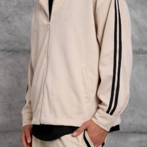 Close view of a beige hooded jacket showing its silver zipper, front pocket, and double black sleeve stripes.