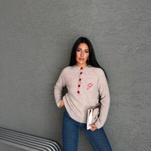 Model in beigem Melange-Strickpullover mit roter Knopfleiste und eingesticktem Initial; sie hält ein Magazin an der Seite.