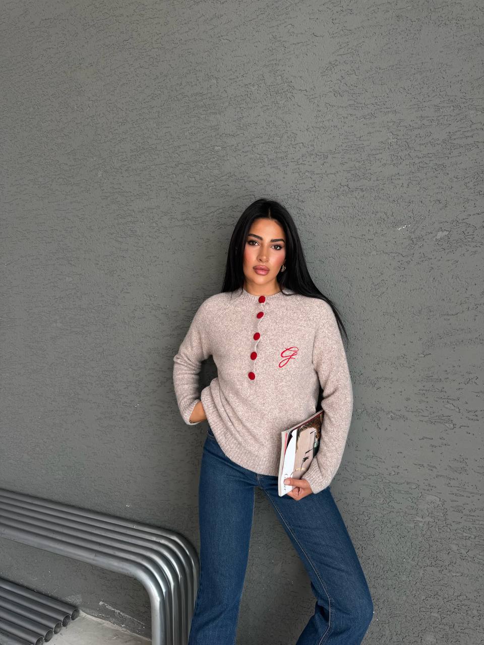 beiger-strickpullover-model-8097 – Türkei-Großhandel Model in beigem Melange-Strickpullover mit roter Knopfleiste und eingesticktem Initial; sie hält ein Magazin an der Seite.