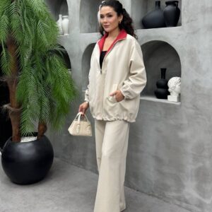 Leicht cremefarbene Steinfarbene Reißverschluss-Jacke und passende gerade Hose, Model hält eine kleine cremefarbene Handtasche seitlich.