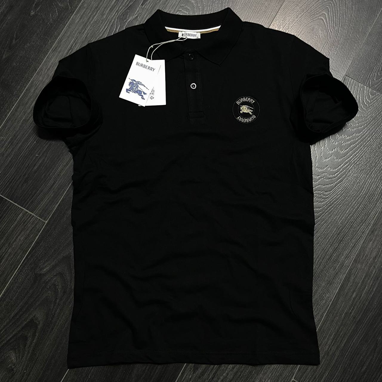 black-polo-crest-4827 Front view of a black collared polo shirt with a button placket and circular embroidered chest crest. available for wholesale in Turkey.