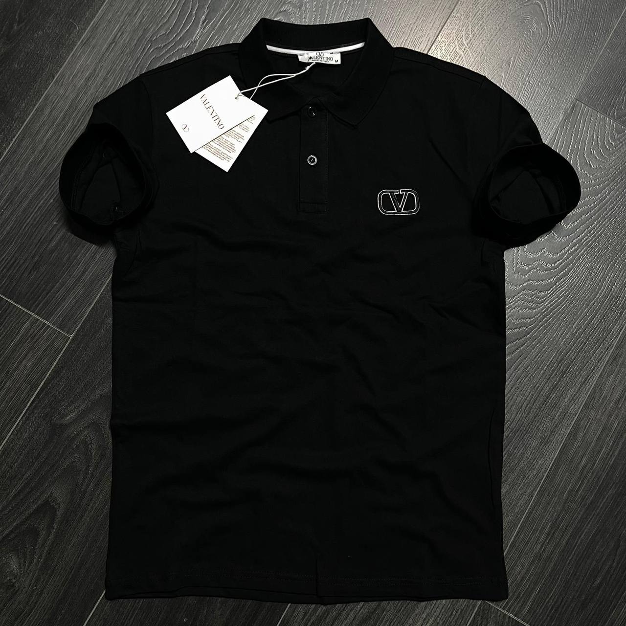 schwarzes-polo-gesticktes-emblem-4827 - Großhandel Türkei Flache Lay-Ansicht eines schwarzen Polohemds mit Kragen, Knopfleiste und umrissenem Brustemblem. Im Großhandel in der Türkei erhältlich - Großhandel Türkei.