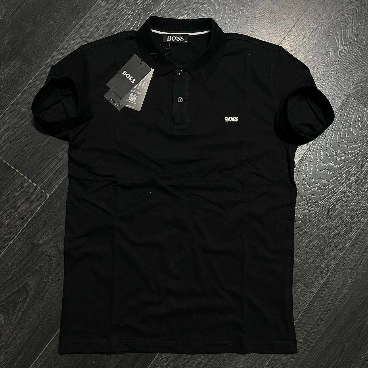 schwarzes-polohemd-vorne-2746 – Türkei Großhandel Schwarzes Kurzarm-Polohemd flach liegend, mit spitzem Kragen, zweiknopf-Knopfleiste und kleinem hellen Brustemblem. Großhandel in der Türkei erhältlich - Türkei Großhandel