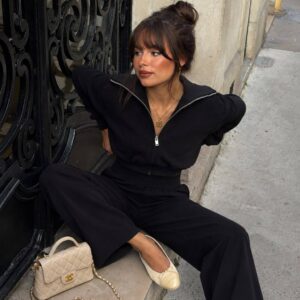 Seated model wearing a black zip jacket with ribbed cuffs and wide-leg trousers.