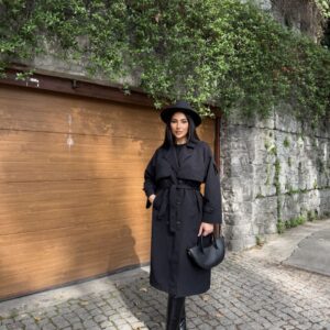 Long black double-breasted trench coat with broad lapels, a tied waist belt, and buttoned cuff straps.