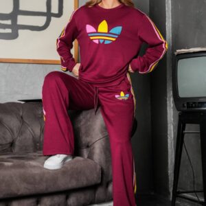 Model seated in a burgundy sweatshirt and loose pants with colorful sleeve and leg stripes and a geometric chest emblem.