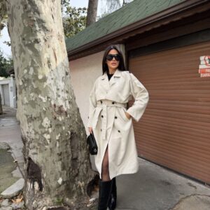 Full-length ivory trench coat with double-breasted buttons, broad lapels, a waist belt, and cuff tabs.